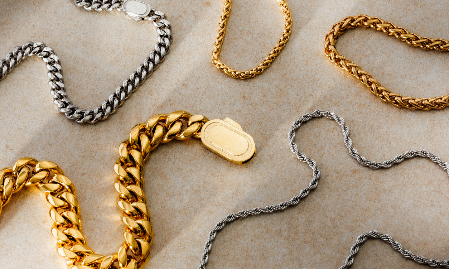 Flat lay of gold and silver CRAFTD chains on a neutral stone background, featuring a mix of Cuban links, rope chains, and sleek minimal styles.