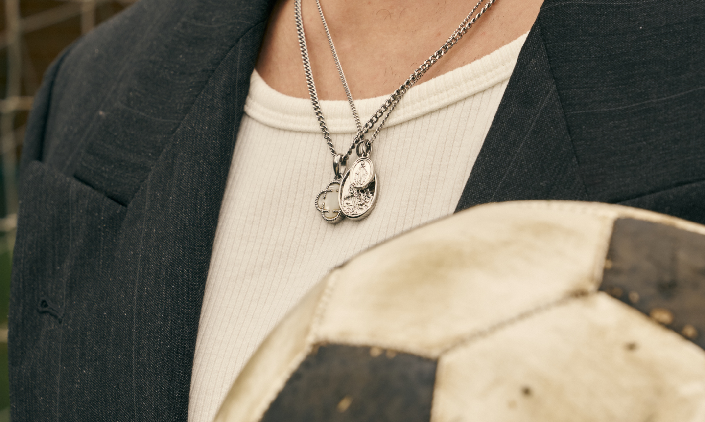 Layered men's silver pendant necklaces styled with a white tee and dark blazer, beside a vintage football for the CRAFTD Summer Lookbook