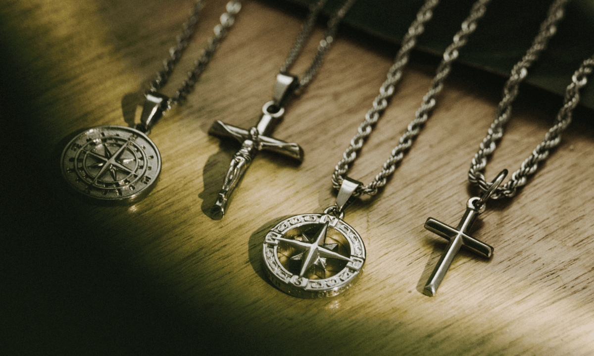Four men’s silver pendants laid on a wooden surface, including two cross designs and two compass designs, each suspended from sterling silver chains. The lighting highlights the polished finish and rugged detail of each piece.